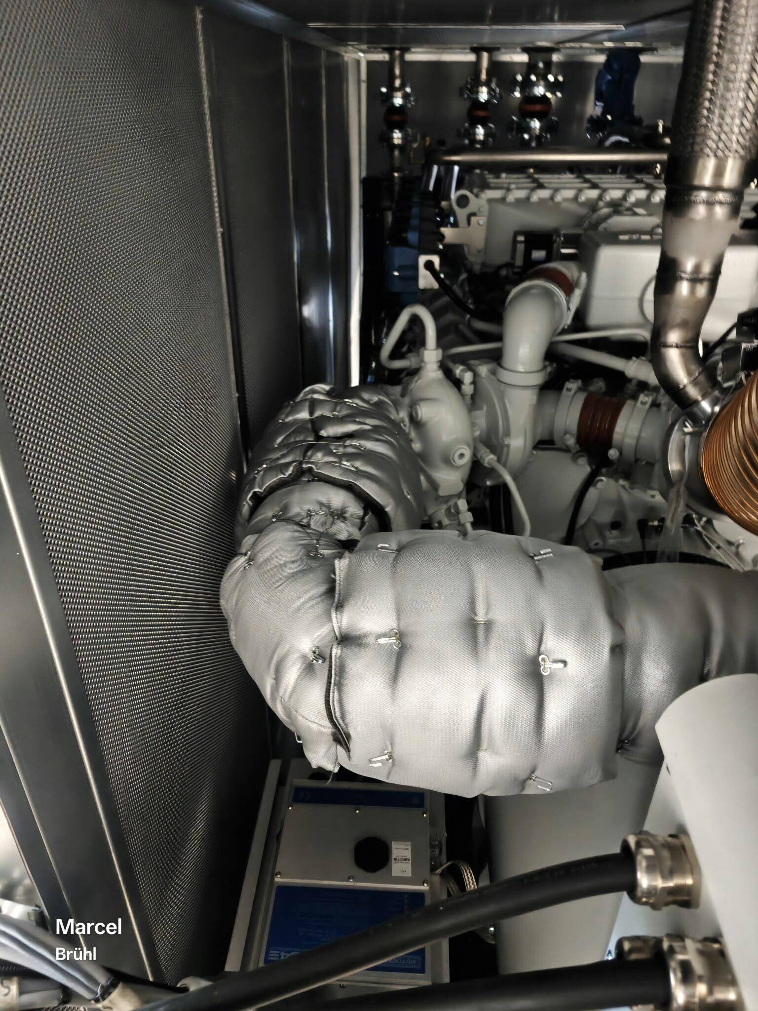 Grijze isolatiematrassen met haken en binddraad rond de uitlaat van een motor in een installatieruimte
