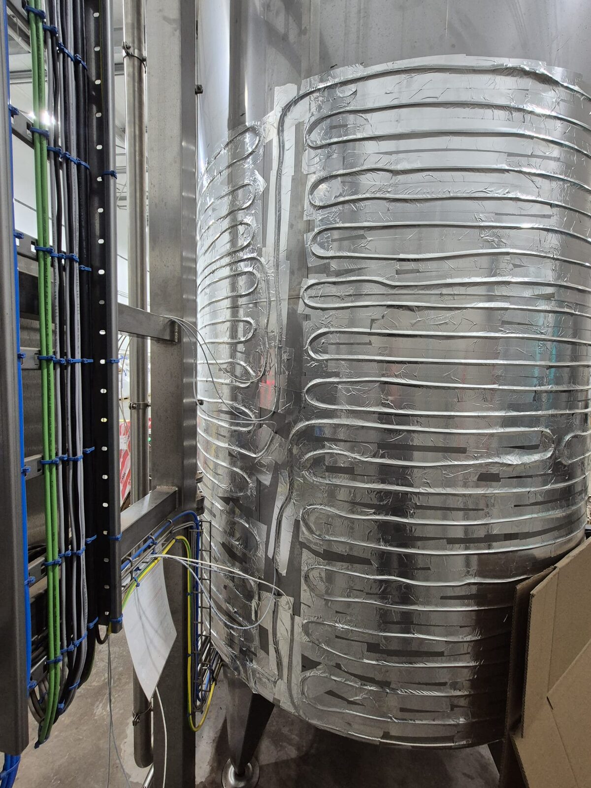 Tank met tracingleidingen in meanderpatroon onder aluminium tape, naast een kast met bekabeling